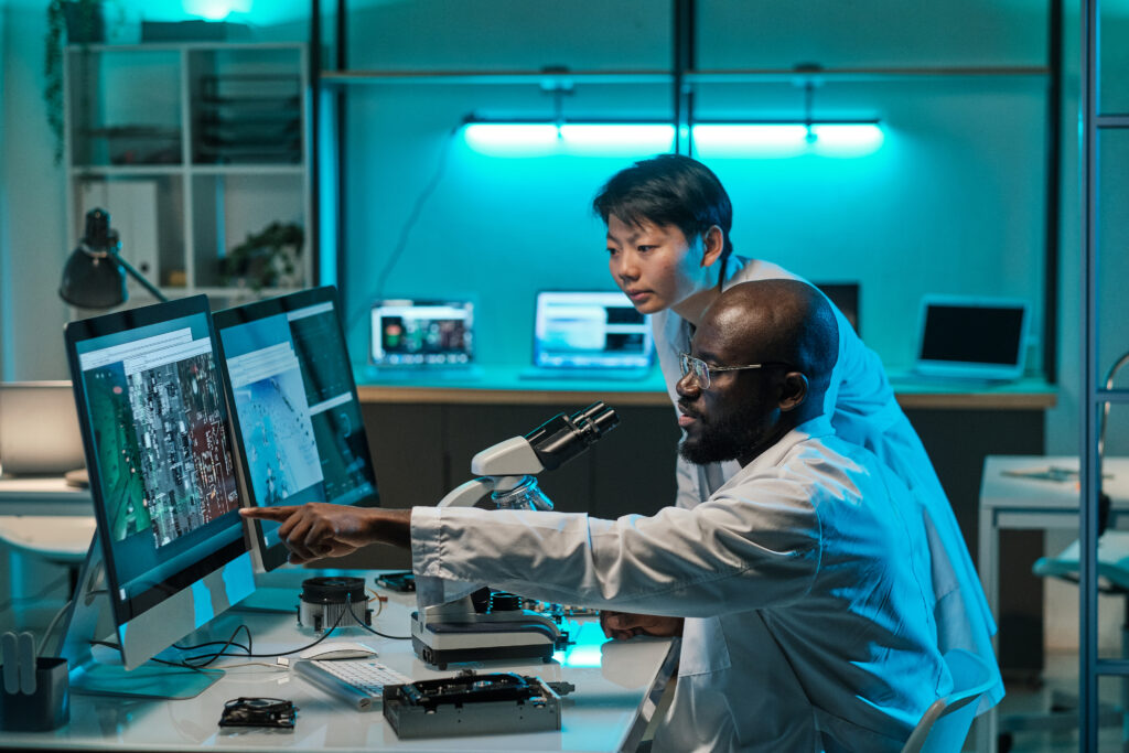 Two young intercultural professionals in whitecoats discussing microchip scheme while one of them pointing at computer screen