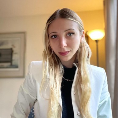 Photo de Rebecca aux cheveux longs et clairs, portant une veste blanche sur un haut sombre. Elle est à l'intérieur, et on aperçoit une lampe sur pied et un tableau encadré en arrière-plan, créant une atmosphère chaleureuse et légèrement éclairée.