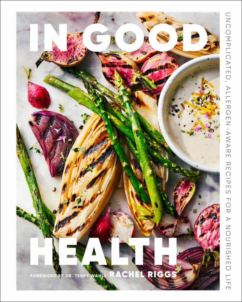 This image is the cover of a book titled "In Good Health" by Rachel Riggs. It features a colorful arrangement of grilled vegetables, including asparagus, onions, and radishes, along with a bowl of dipping sauce.