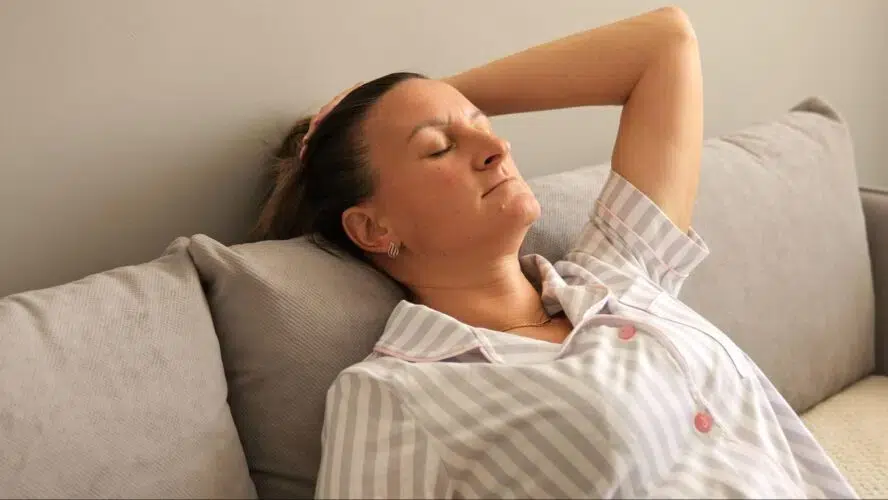 L'image montre une femme atteinte d'EM/SFC se reposant sur un canapé. Elle porte un pyjama rayé et se penche en arrière, les yeux fermés.