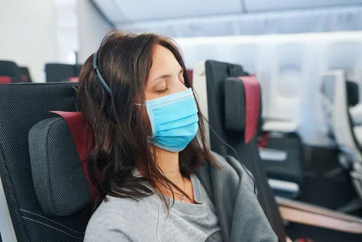 A woman wearing a medical face mask and headphones is resting with her eyes closed on an airplane. She is wrapped in a blanket.